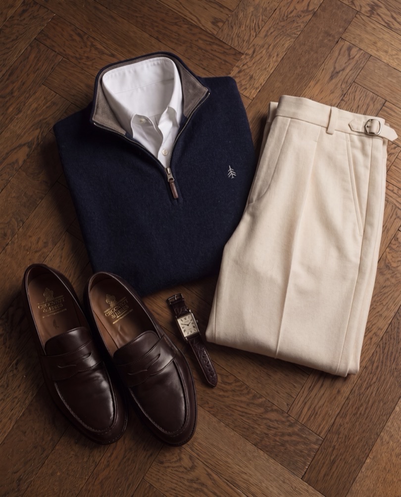 A navy quarter-zip folded with cream trousers, brown loafers and a watch on a herringbone floor.
