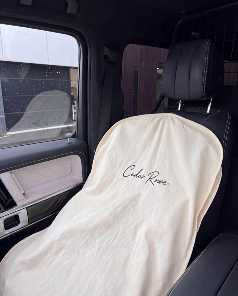 A cream Cedar Rowe seat cover, signed with the script wordmark, fitted over a dark leather car seat.